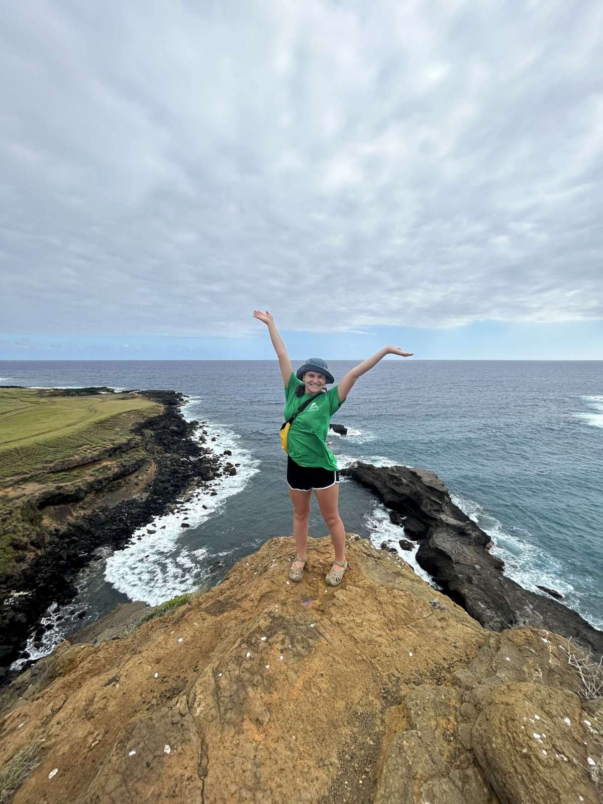 image shows a female student posing with raised arms and the ocean behind her