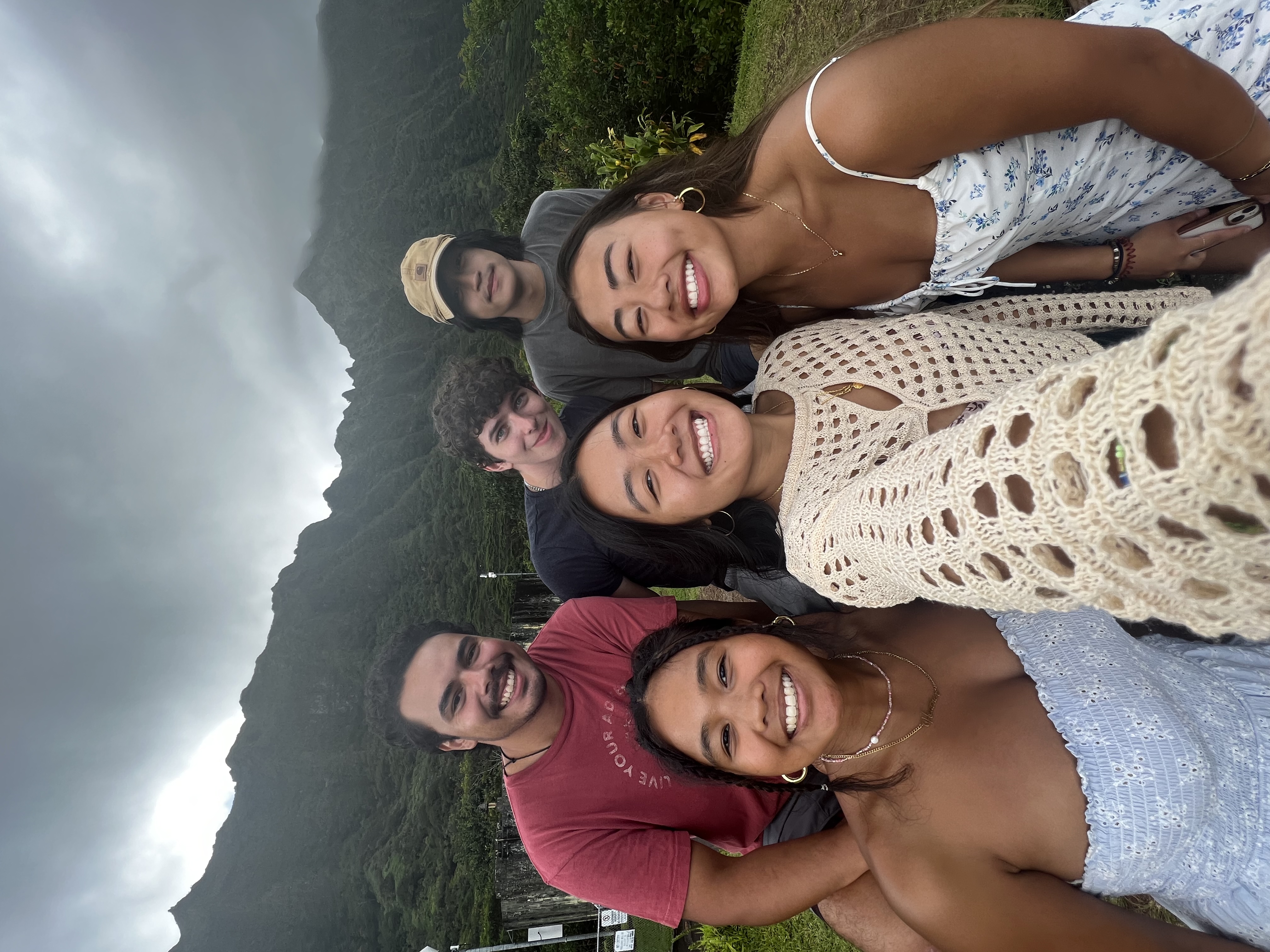 Image shows a group of students posing in a "selfie" style. There are mountains in the background against a cloudy sky.