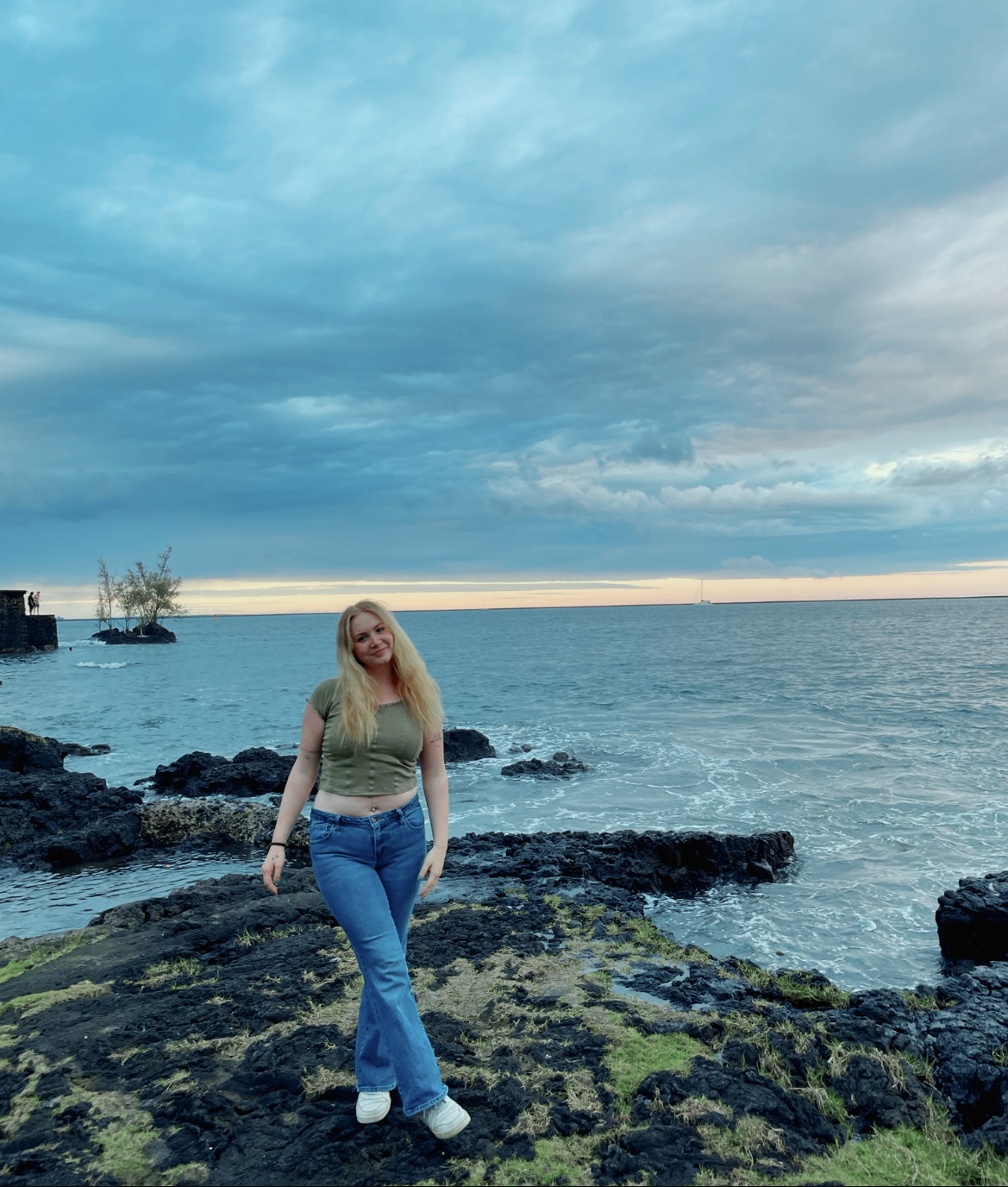 Image shows a figure posing in front of a rocky coastline with a cloudy background